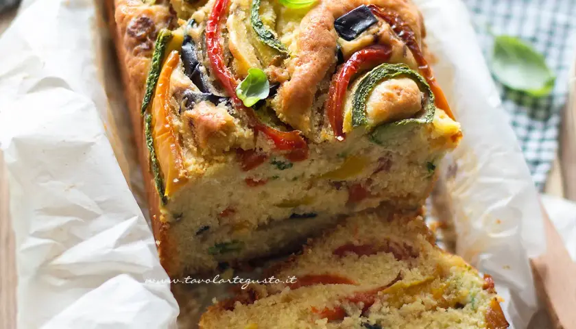 Plumcake salato ricco di verdure colorate come zucchine, peperoni e olive, guarnito con basilico fresco.
