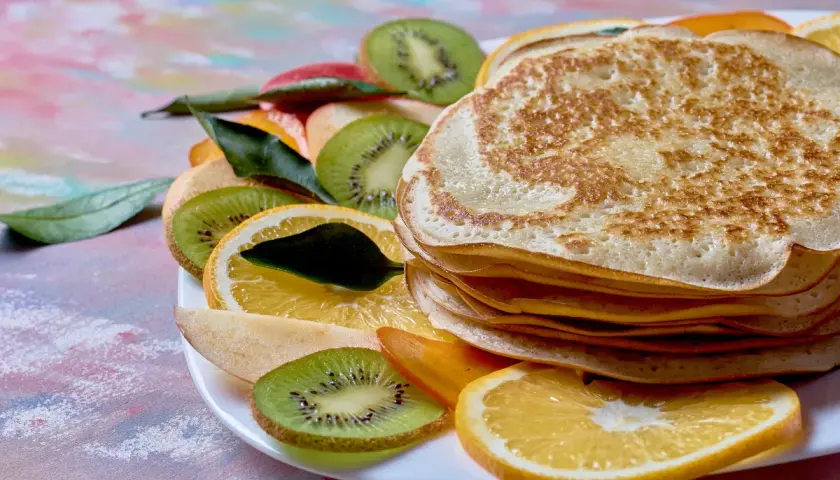 Stos naleśników z budyniem, otoczony plasterkami kiwi, pomarańczy i jabłka, ozdobiony listkami.