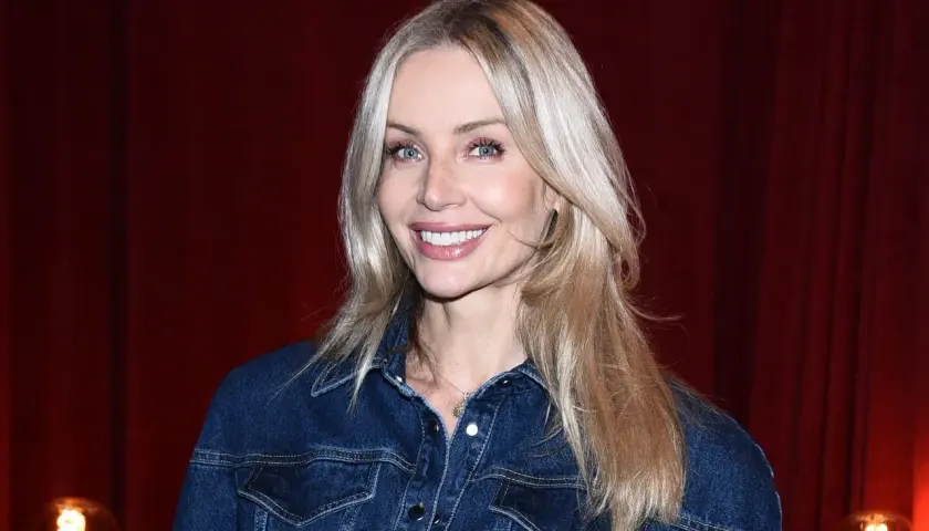 Uśmiechnięta blondynka z francuską grzywką w dżinsowej kurtce na tle czerwonej kurtyny.