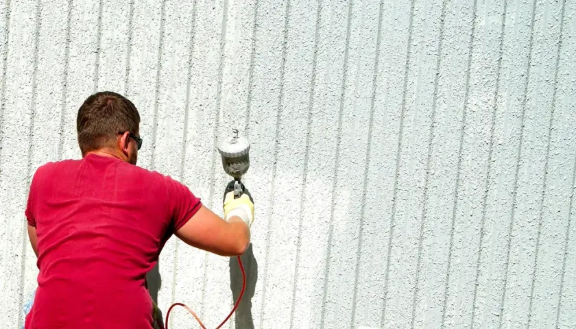Uomo con maglietta rossa e occhiali da sole sta pitturando casa a spruzzo con una pistola.