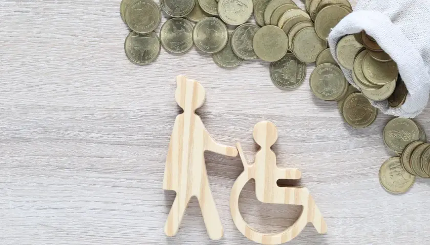 Drewniane figurki osoby pomagającej osobie na wózku inwalidzkim i monety. Czy zasiłek pielęgnacyjny wlicza się do dochodu?