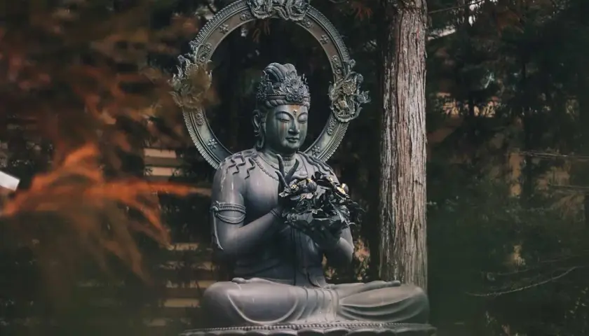 Kamienna statua Buddy w pozycji medytacyjnej, otoczona drzewami. Symbol spokoju i buddyzmu.