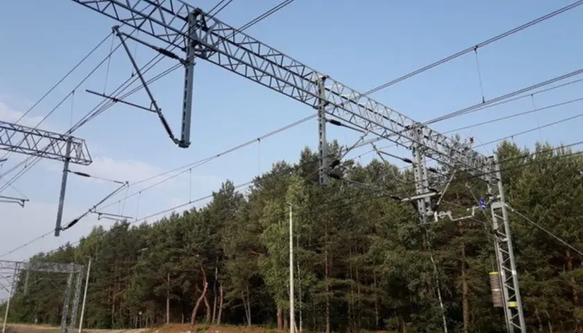 Trwa budowa sieci trakcyjnej nad torami kolejowymi, otoczona gęstym lasem sosnowym.