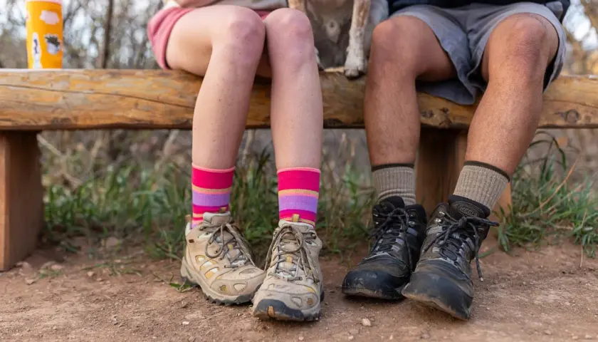 Colorful striped and neutral socks to wear with hiking boots, worn by two people sitting on a wooden bench with hiking boots on.