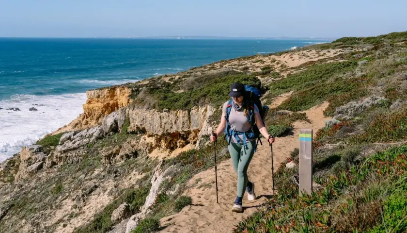 Wanderin auf dem Fisherman's Trail in Portugal. Eine Frau mit Rucksack und Stöcken wandert auf einem sandigen Küstenpfad mit Blick auf das Meer.