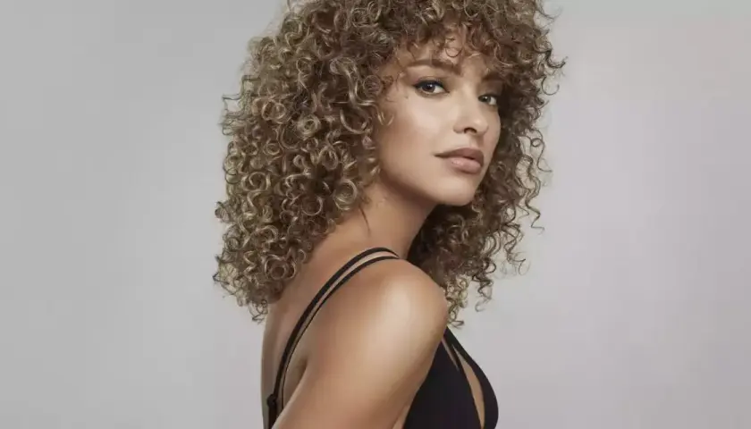 Kobieta z kręconymi, blond grzywkami i w czarnej sukience na jasnym tle.