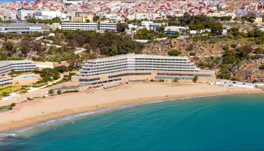 Widok na hotel przy piaszczystej plaży i turkusowym morzu, z miastem w tle.