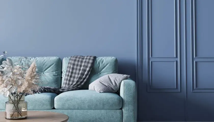 Miętowa kanapa z kocem i poduszką na tle ścian w kolorze baby blue. To znaczy spokój i świeżość.