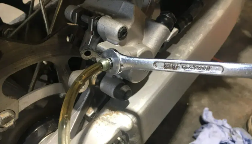 Klucz nasadowy odkręca odpowietrznik podczas wymiany płynu hamulcowego w motocyklu. Widać tarczę hamulcową i zacisk.