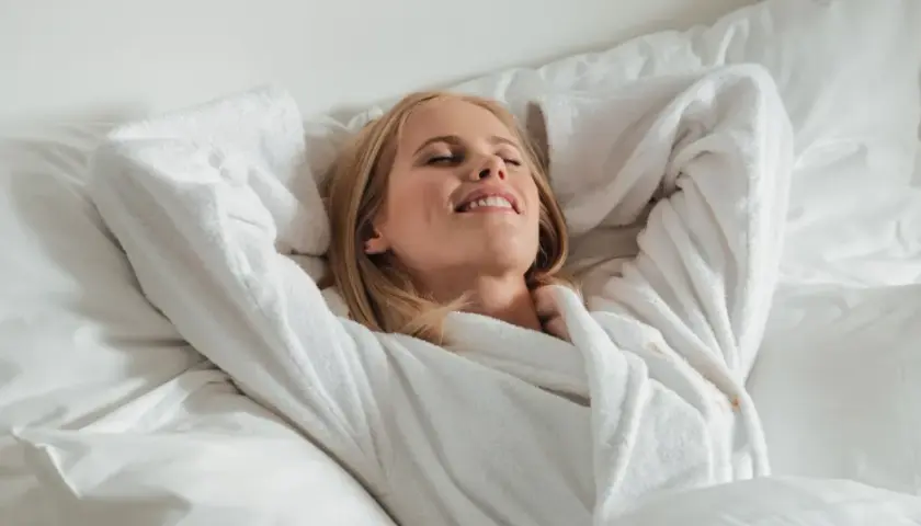 Frau entspannt im Bademantel im Bett, genießt den Moment. Vielleicht denkt sie über die richtige 5-HTP Dosierung nach, um sich noch besser zu fühlen.