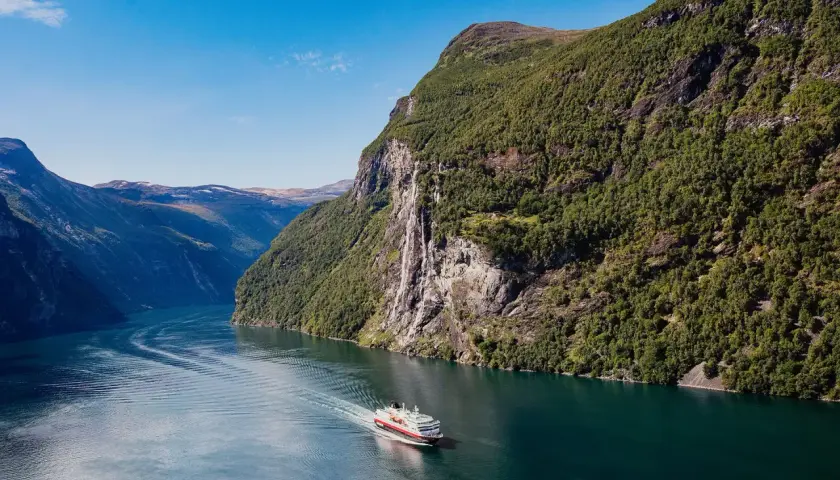 Ein Schiff fährt durch einen norwegischen Fjord im August. Das Wetter ist sonnig, die Berge sind grün und ein Wasserfall stürzt herab.