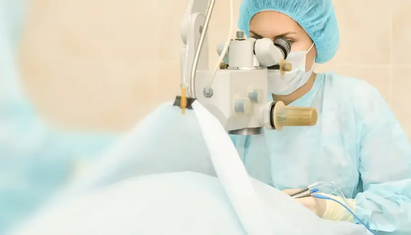 Chirurg w masce i czepku wykonuje operację zaćmy, używając mikroskopu i precyzyjnych narzędzi.