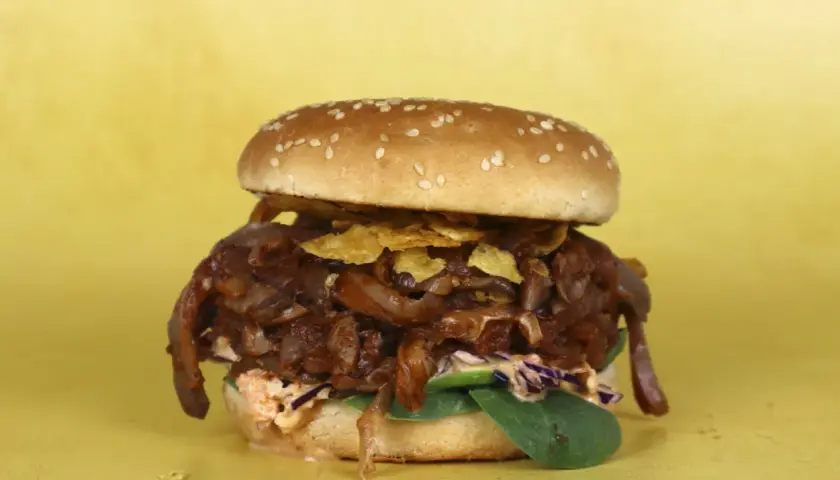 Hamburguesa con pulled pork vegano, cebolla caramelizada y crujientes patatas fritas sobre fondo amarillo.