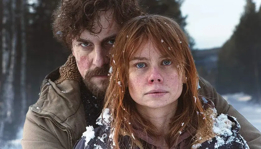 Julia Schlaepfer och en man med skägg och lockigt hår, båda med snö i håret, ser allvarligt in i kameran i en vinterlig skogsmiljö.