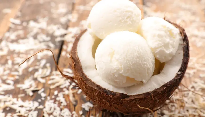 Tre palline di cremoso gelato al cocco servite in una noce di cocco su un tavolo di legno con scaglie di cocco sparse.