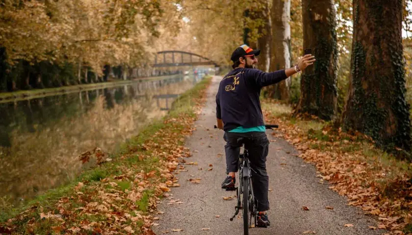 Hombre en bicicleta toma selfie en el Canal du Midi, rodeado de árboles otoñales y hojas caídas.