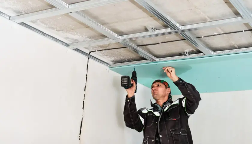 Un ouvrier réalise la pose de suspente et fourrure pour un faux plafond en plaques de plâtre bleu.