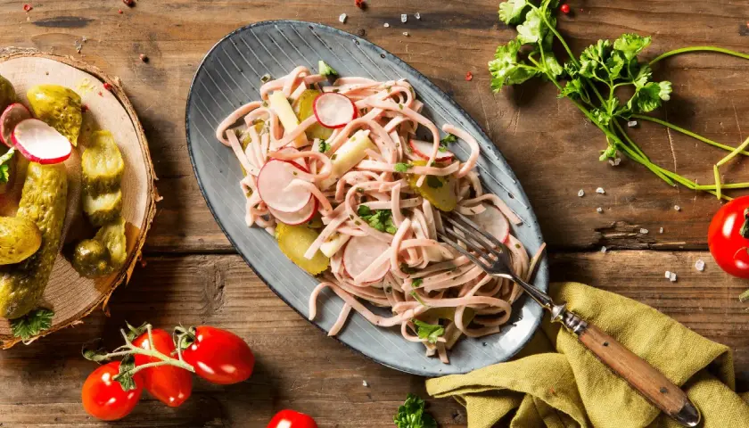 Ein köstlicher low carb picknick Teller mit Wurstsalat, Radieschen, Gewürzgurken und Tomaten auf rustikalem Holztisch.