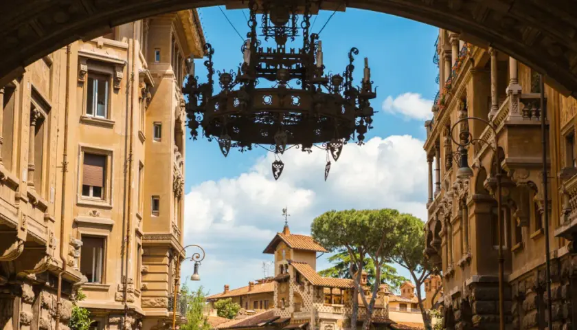 Un imponente lampadario domina la vista del quartiere Coppedè, incorniciato da palazzi storici e un cielo azzurro con nuvole bianche.