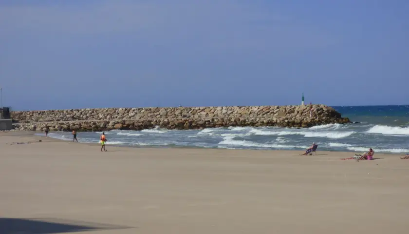 Una playa tranquila con una rompeolas de rocas. Gente relajándose en sillas de playa y paseando. El mar azul y el cielo despejado crean una escena de verano perfecta.