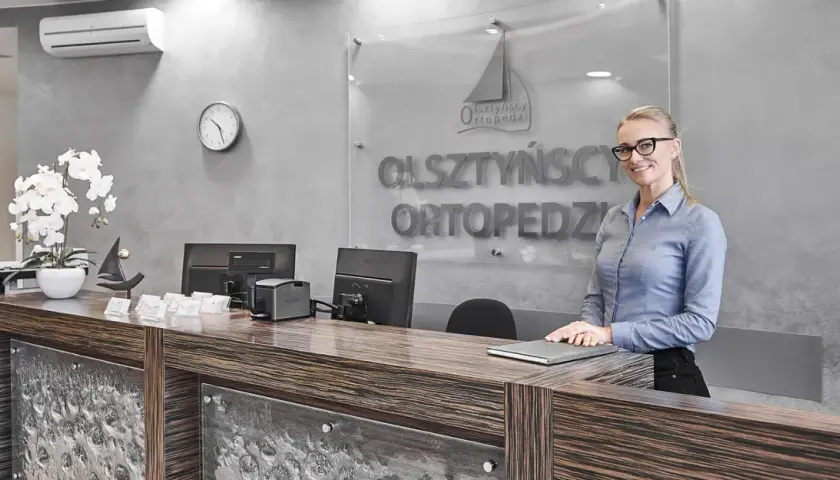 Recepcjonistka w okularach stoi przy ladzie w nowoczesnej klinice ortopedycznej.