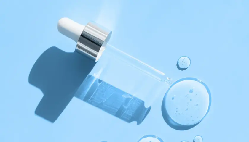En pipett med klar vätska, som innehåller hyaluronsyra och retinol, ligger på en ljusblå bakgrund med droppar av serum.