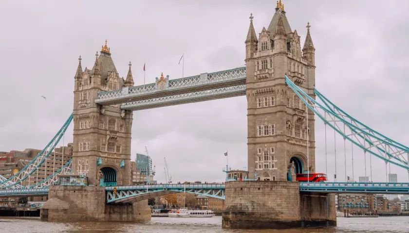 Tower Bridge w Londynie, symbol miasta, opowiada swoją historię. Czerwony autobus przejeżdża przez most, a w tle widać nowoczesne budynki.
