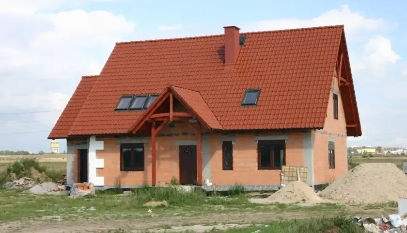 Obecny sposób zagospodarowania terenu inwestycji to budowa domu z czerwonym dachem, otoczonego materiałami budowlanymi i zielenią.