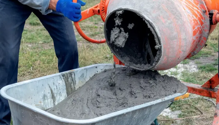 Béton frais coulant d'une bétonnière orange dans une brouette. Un ouvrier en gants bleus manipule la manivelle pour déverser le mortier béton.