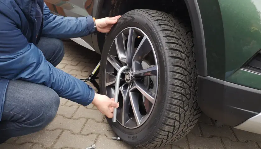 Osoba w niebieskiej kurtce i jeansach dokonuje wymiany koła w samochodzie, używając podnośnika i klucza.