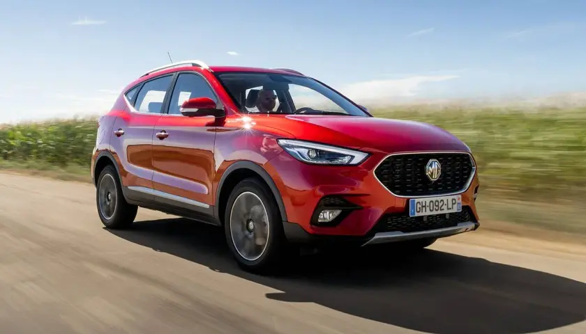 MG ZS en prueba, rojo brillante, circulando por un camino rural con campos verdes y cielo azul.