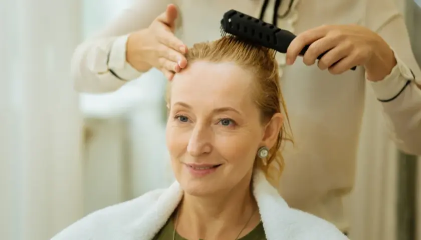 Stylist czesze blondynce cienkie i rzadkie włosy, przygotowując ją do nowej fryzury.