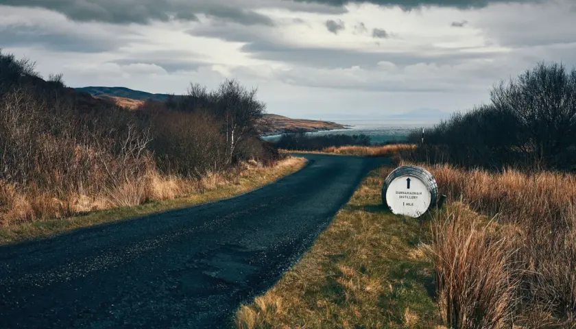 Route sinueuse menant à la distillerie Bunnahabhain, avec un fût indiquant la direction. Paysage écossais sauvage sous un ciel menaçant.