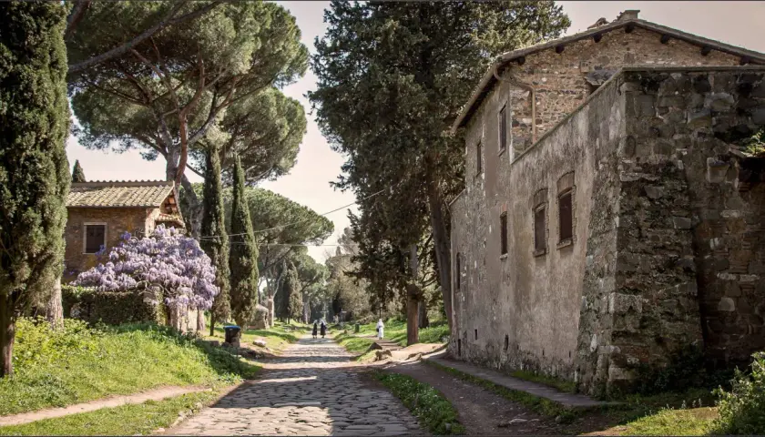 Une ancienne voie romaine à Rome, bordée de cyprès et de pins parasols, avec des maisons en pierre et des glycines en fleurs.