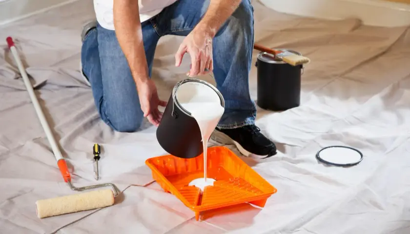 Uomo versa vernice bianca in una vaschetta arancione, pronto per pitturare soffitto con pennello. Attrezzi da pittura a portata di mano.