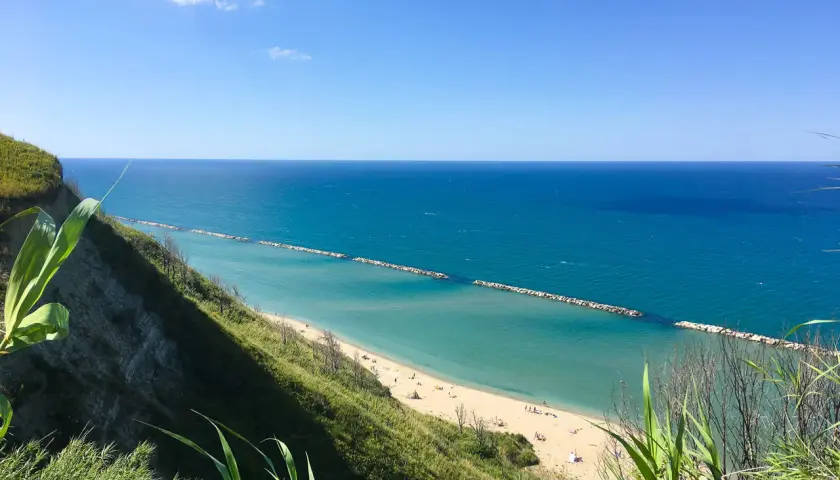 Vista panoramica della spiaggia di Pesaro con mare azzurro e scogliera verde. La focara è un ricordo lontano, ma la bellezza del paesaggio resta.