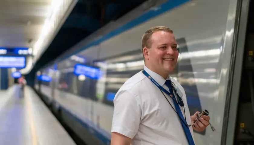 Uśmiechnięty mężczyzna w białej koszuli i granatowym krawacie, z identyfikatorem PKP Intercity, trzyma w dłoni narzędzie. To część drużyny konduktorskiej gotowej do podróży.