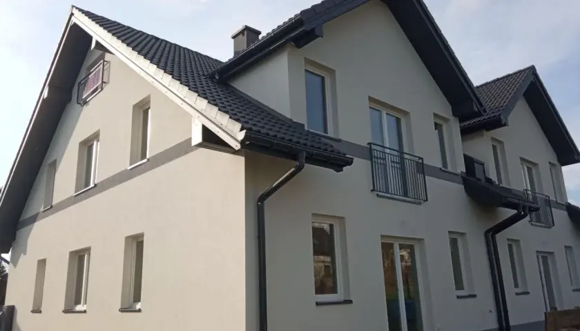 Nowy dom z jasną elewacją i ciemnym dachem. Wokół zielona rząska trawy, a na ścianach liczne okna i balkony.