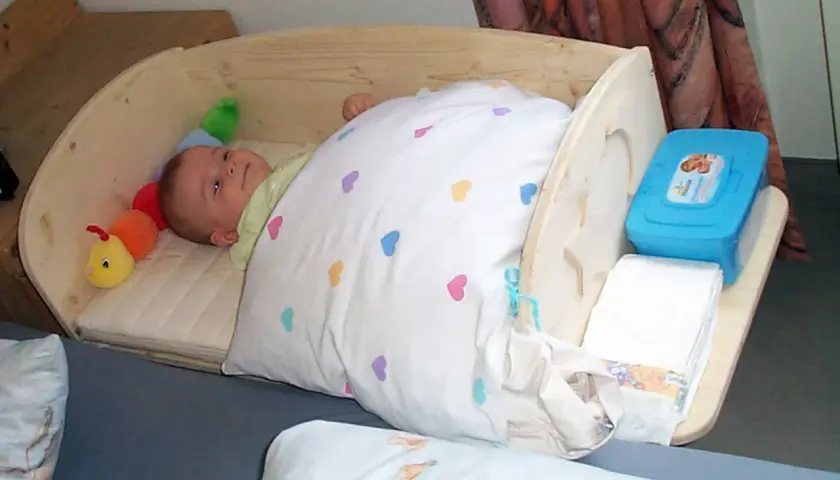Ein süßes Baby schläft in einem selbstgebauten Babybett aus Holz. Neben ihm liegt ein bunter Raupen-Greifling. Windeln und Feuchttücher sind griffbereit.