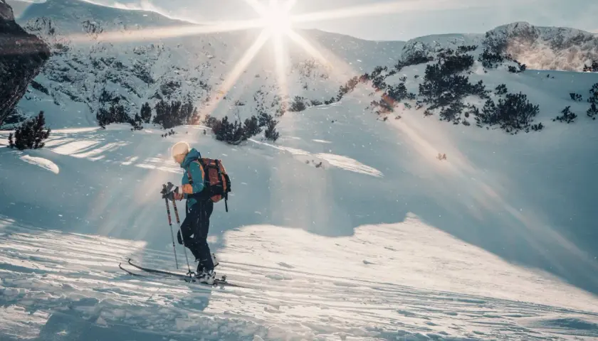 Skitouring co to? Narciarz z plecakiem wspina się po ośnieżonym stoku, słońce przebija się przez chmury, tworząc magiczną atmosferę.