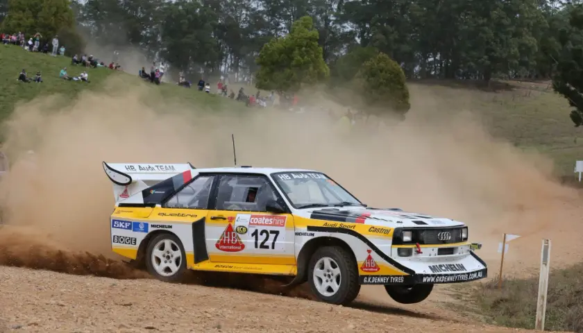 Klasyczny Audi Quattro z grupy B, oznaczony numerem 127, pędzi po szutrowej drodze, wzbijając tumany kurzu.