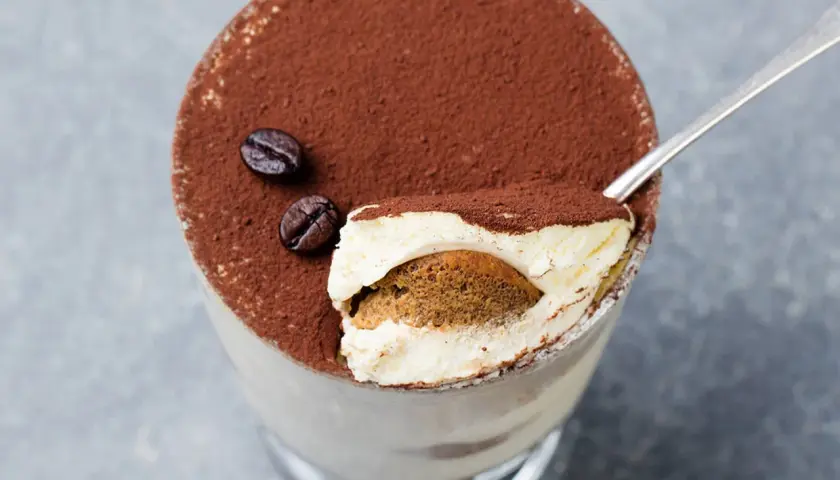 Un goloso tiramisù giallo zafferano, cosparso di cacao e chicchi di caffè, pronto per essere gustato con un cucchiaino.