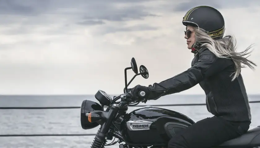 Mujer rubia en moto Triumph negra, con casco y chaqueta de cuero, disfrutando del paisaje. Las motos Triumph se fabrican con pasión.