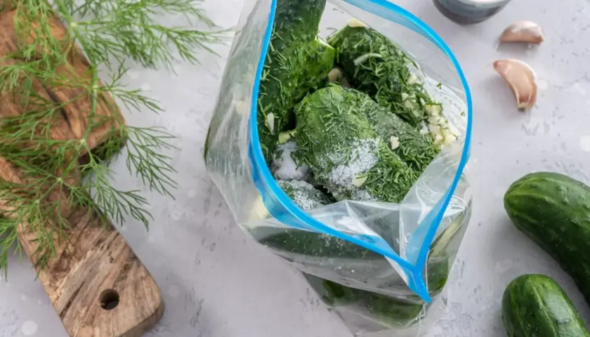 Przygotowanie ogórków małosolnych bez wody. W przezroczystej torbie zamykanej na zamek błyskawiczny znajdują się świeże ogórki, koperek i czosnek, posypane solą.