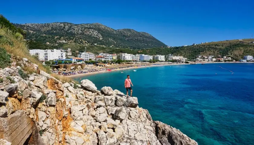 Mann steht auf Felsen mit Blick auf eine Bucht mit Strand und Hotels. Perfekt, um zu wissen, wo in Albanien Urlaub machen!