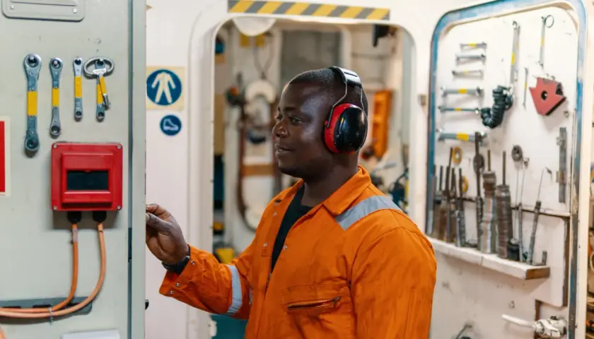 Técnico con auriculares rojos ajusta un panel de control. Herramientas cuelgan en la pared. El nivel de ruido podría superar la escala db.