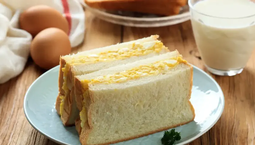 Un délicieux sandwich aux œufs, garni de pain blanc moelleux, servi sur une assiette bleue avec un verre de lait.