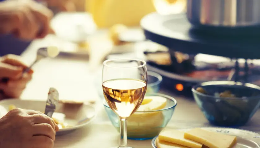 Un verre de vin blanc sec ou moelleux accompagne une raclette conviviale. Des morceaux de fromage et des cornichons sont prêts à être dégustés.