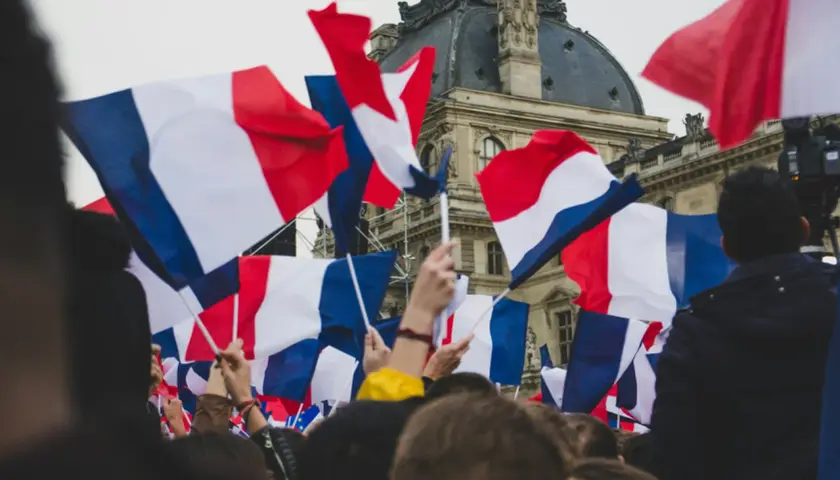Tłum ludzi trzyma flagi francuskie. Widać charakterystyczne biało-niebiesko-czerwone pasy, jak wygląda flaga francuska.