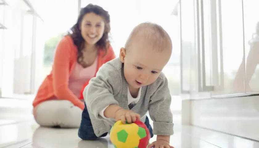 Dziecko raczkuje, ćwicząc motorykę dużą co to jest, sięgając po kolorową piłkę. Uśmiechnięta mama obserwuje.
